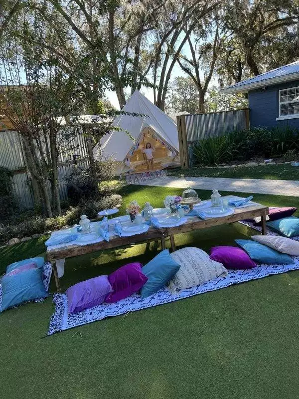 Outdoor setting featuring a low wooden table with blue and white tableware, surrounded by colorful cushions and rugs, adjacent to a teepee tent perfect for a slumber party in the grassy yard.