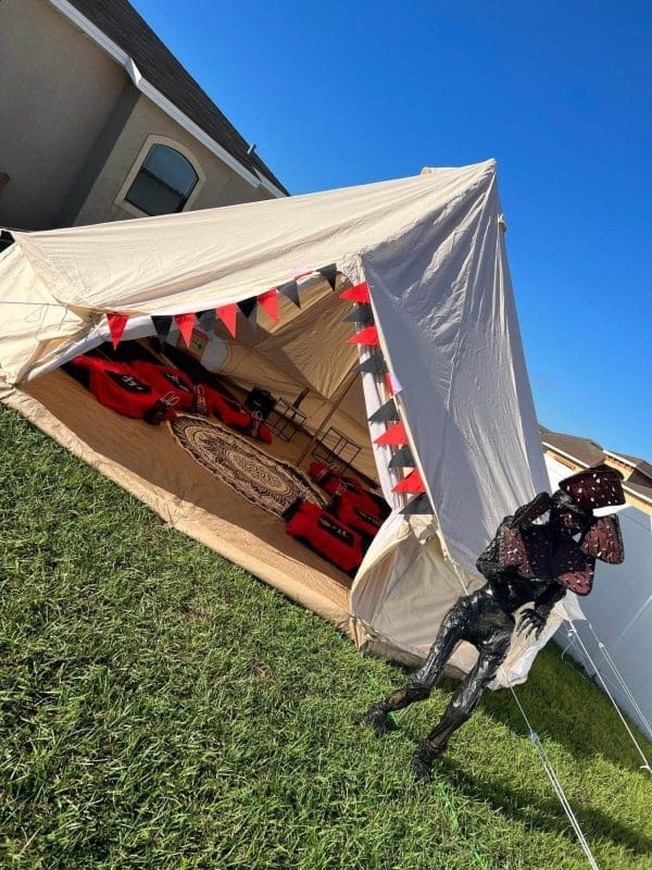 A large beige tent with an open front is set up on a grassy lawn, perfect for a kids party rental. Inside, red and black decorative elements complement the circular rug, while a dinosaur figure stands guard near the entrance of the tent.