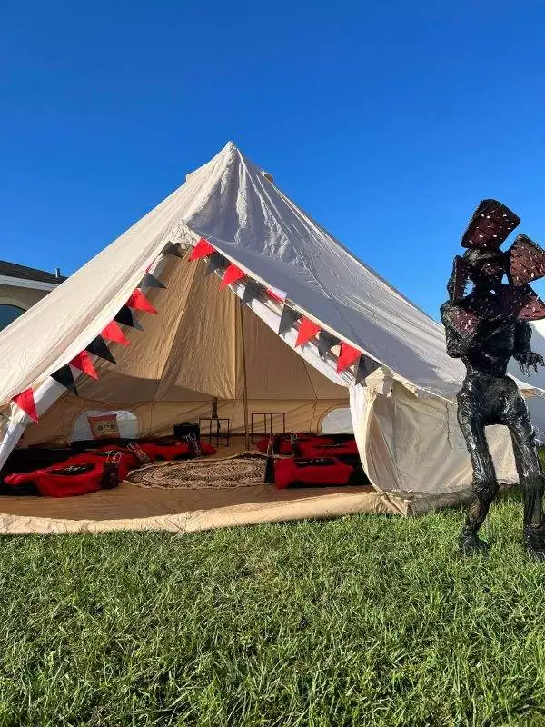 A large canvas tent with an open front, decorated with red and black bunting, is set up on grass. Inside are red sleeping bags and pillows, perfect for a slumber party. A tall, dark creature statue stands nearby. This setup can be arranged through our premium party rental service.