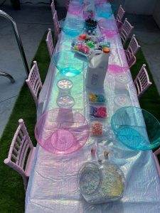 A long table with transparent plastic bowls, a large container of glue, small jars with colored beads, and other craft supplies is arranged for a children's activity, perfect for any party rental.