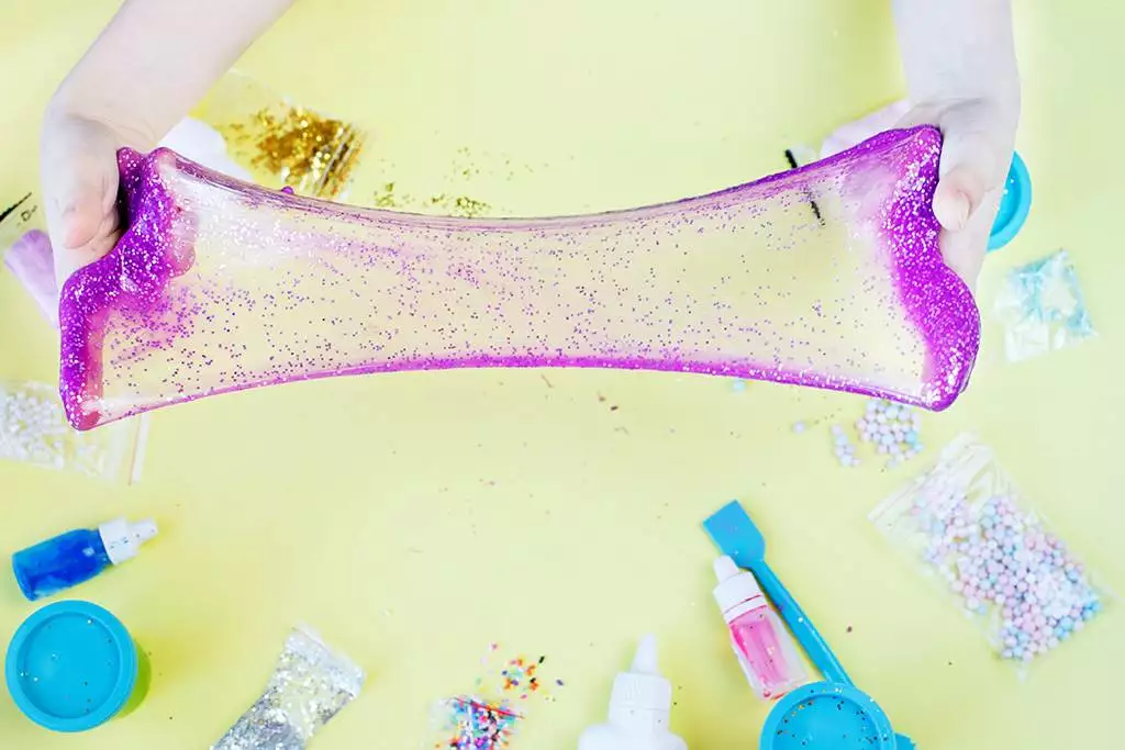 Two hands stretch a piece of pink and purple glittery slime over a table, surrounded by various craft supplies, creating the perfect scene for a kids party rental.