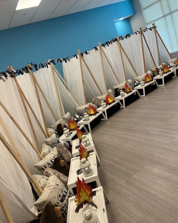 A room is set up with indoor teepee tents along the walls, each accompanied by small tables with faux campfire decorations and pillows, creating a cozy slumber party atmosphere perfect for party rentals.