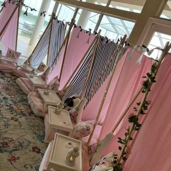 A row of indoor pink and black striped teepees with white cushions and small tables set up for a party rental event. Each table holds a small lantern. The setting overlooks a bright outdoor area through large windows, making it perfect for a memorable party service.