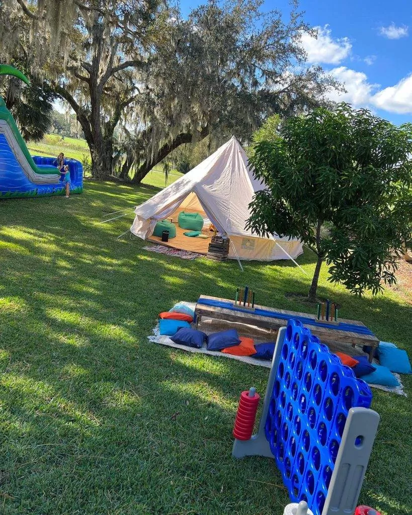 A grass lawn with a white tent, a water slide, and a large Connect Four game awaits. Cushions and a low table are set up near the tent under a tree, perfect for your next kids party rental.