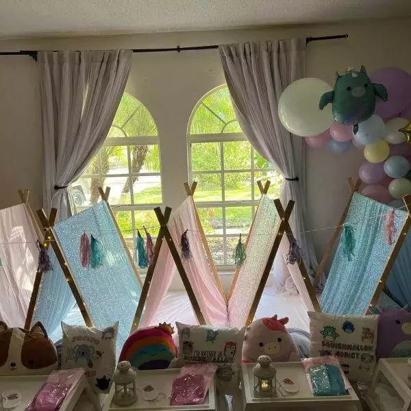 A room setup for a children's sleepover features small tents, colorful decorative pillows, and fairy lights. Perfect for a slumber party, the space is adorned with balloons and curtains framing a large window in the background. Consider this charming scene for your next kids' party rental.