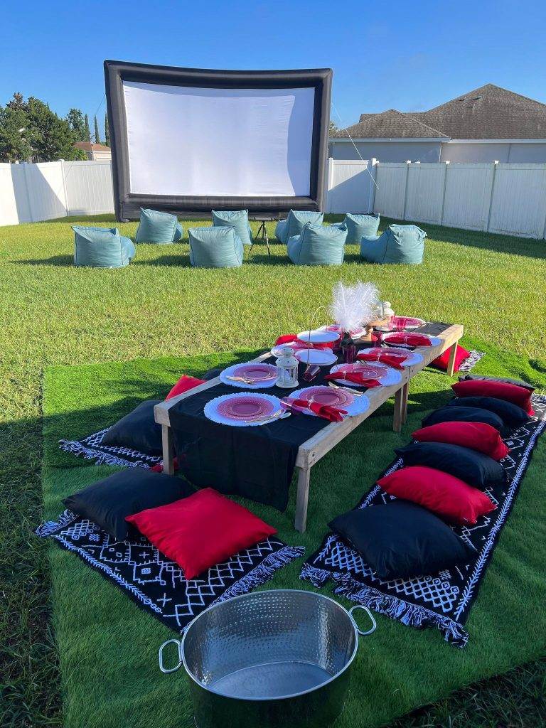 Outdoor movie setup with a large inflatable screen, bean bag seating, and a low picnic table set with plates, candles, and red and black pillows on a grassy area—perfect for a kids party rental. Our party service can even include themed teepees for an extra touch of magic.