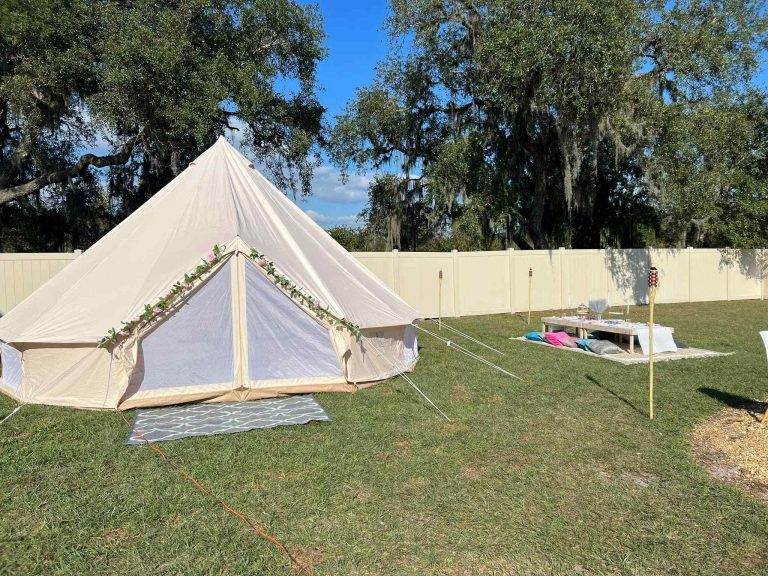 A large canvas tent is set up in a grassy yard beside a wooden table with benches. Tiki torches light up the area, perfect for a lively slumber party. A beige fence lines the perimeter, while trees stand tall in the background. Consider our party service for your next event!