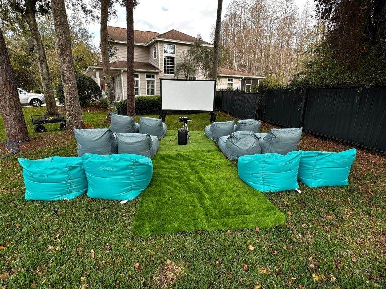 Outdoor seating setup with teal bean bags arranged in rows facing a projector screen in a yard. A house and trees are visible in the background, making it perfect for a cozy party rental experience.