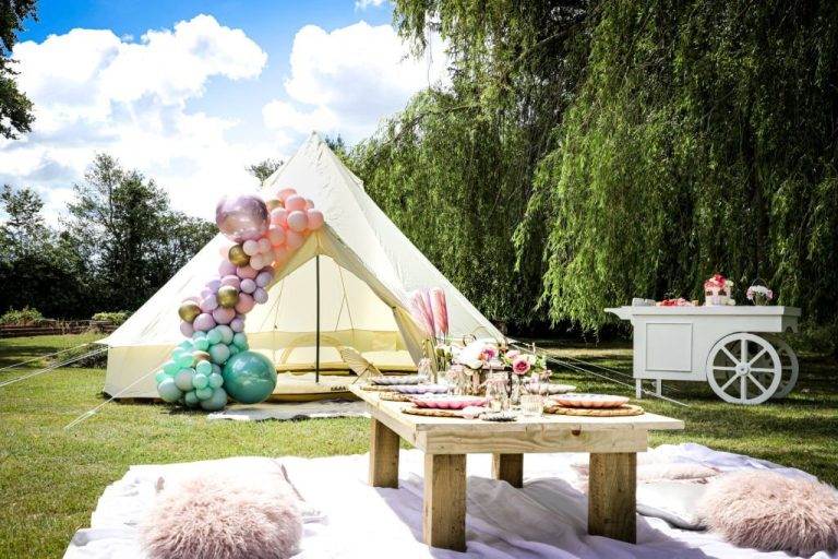 A white tent decorated with balloons stands beside a table set for an outdoor meal near a tree, showcasing the perfect setting for a kids party rental. A cart in the background and pillows on a picnic blanket in the foreground complete this charming scene.