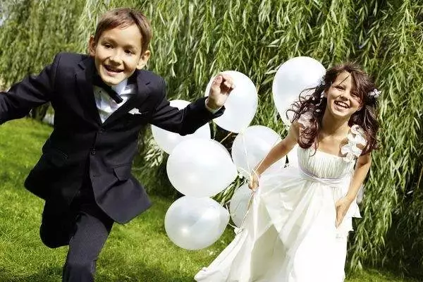 Two children, one in a suit and one in a white dress, happily running outdoors with white balloons in hand on a sunny day amidst greenery, not far from where the teepee for the slumber party is set up.