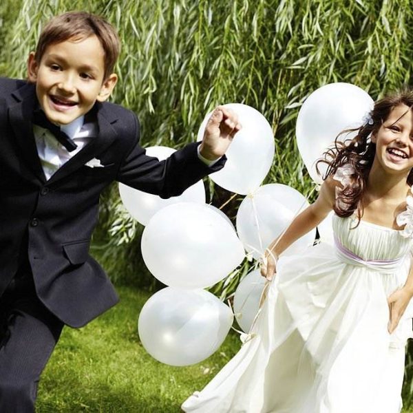 Two children, one in a suit and one in a white dress, happily running outdoors with white balloons in hand on a sunny day amidst greenery, not far from where the teepee for the slumber party is set up.