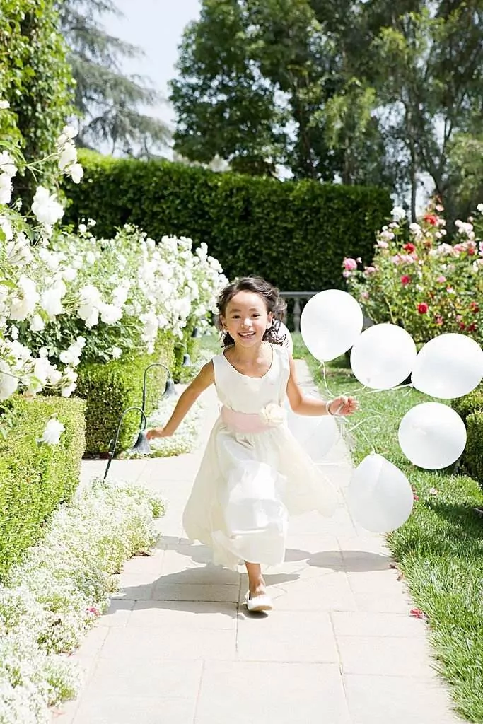 A young girl in a white dress runs along a garden path holding four white balloons. She smiles, surrounded by blooming flowers and green hedges on a sunny day, the perfect setting for her colorful teepee slumber party later that evening.