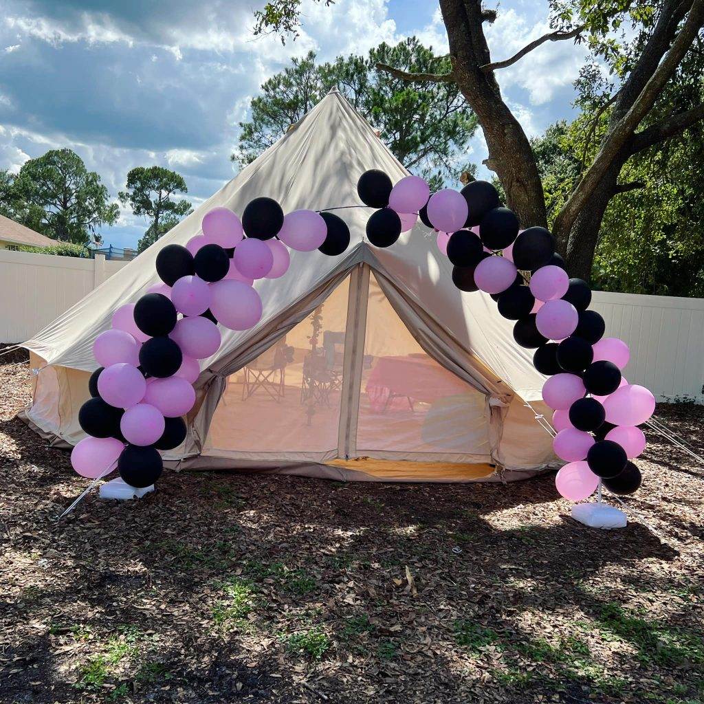 A canvas bell tent decorated with a balloon arch in black and pink, set up outdoors under a clear sky, accessible through an appointment booking.