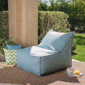 A light blue outdoor lounge chair, perfect for a kids' party rental, is placed on a patterned rug next to a pillar, with green bushes in the background. There are various pillows and a small bowl on the ground beside the chair.