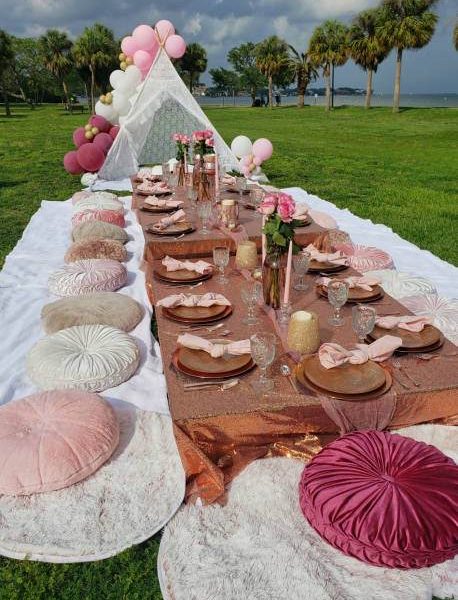 A picnic setup with a long table in a meadow. The table has pink and beige decor, glassware, plates, candles, flower vases, and is surrounded by round cushions. Perfect for a kids party rental or any event needing party service. A small canopy is in the background.