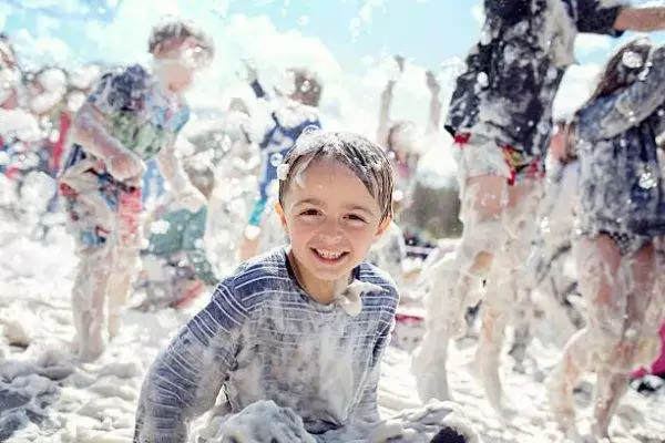 A child smiles while having a foam party sitting in foamy bubbles, surrounded by other children playing in the foam at a fun slime party.