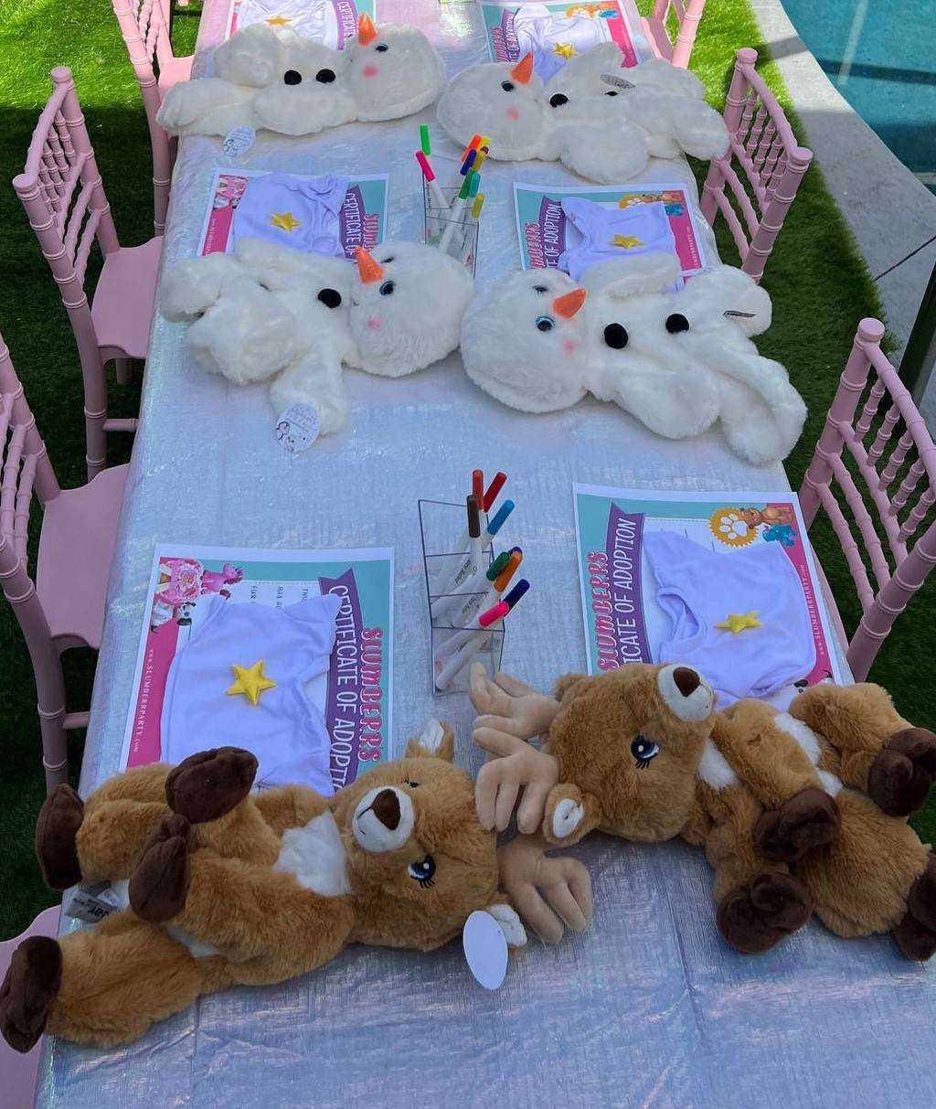 A table adorned with activity sheets, markers, plush reindeer toys, and snowman face pillows set up for a children's event. Pink chairs surround the table, making it ideal for a kids party rental.