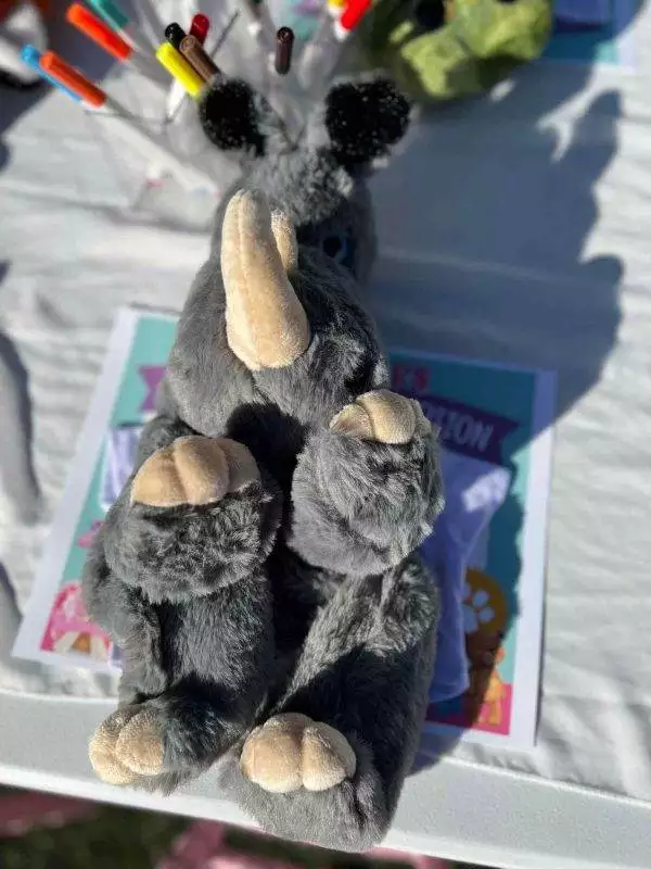 A gray stuffed animal with large ears and black tufts on its head is lying on a table, surrounded by colorful markers and other items, ideal for a kids party rental.