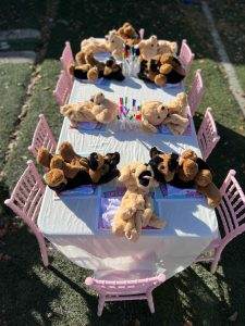 Stuffed teddy bears sitting around a small table set with crayons and paper, outdoors on a sunny day, ready for a build-a-bear party.