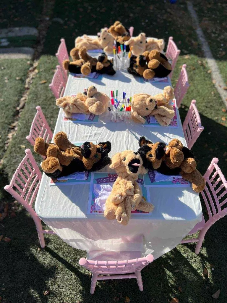 Teddy bears seated around two small pink tables set for a Build A Stuffed Animal party outdoors, with coloring books and crayons.