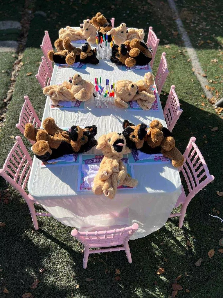 Outdoor children's Build A Stuffed Animal party table set up with stuffed teddy bears seated around, under trees casting shadows, with straws and napkins ready.