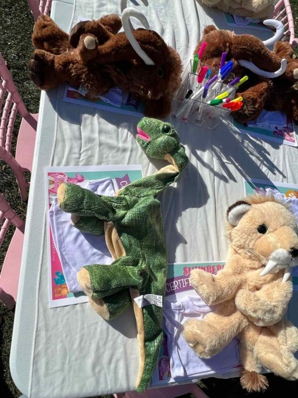 Stuffed animals and pens displayed on a table at a Build A Stuffed Animal party, including a green dinosaur and a brown teddy bear.