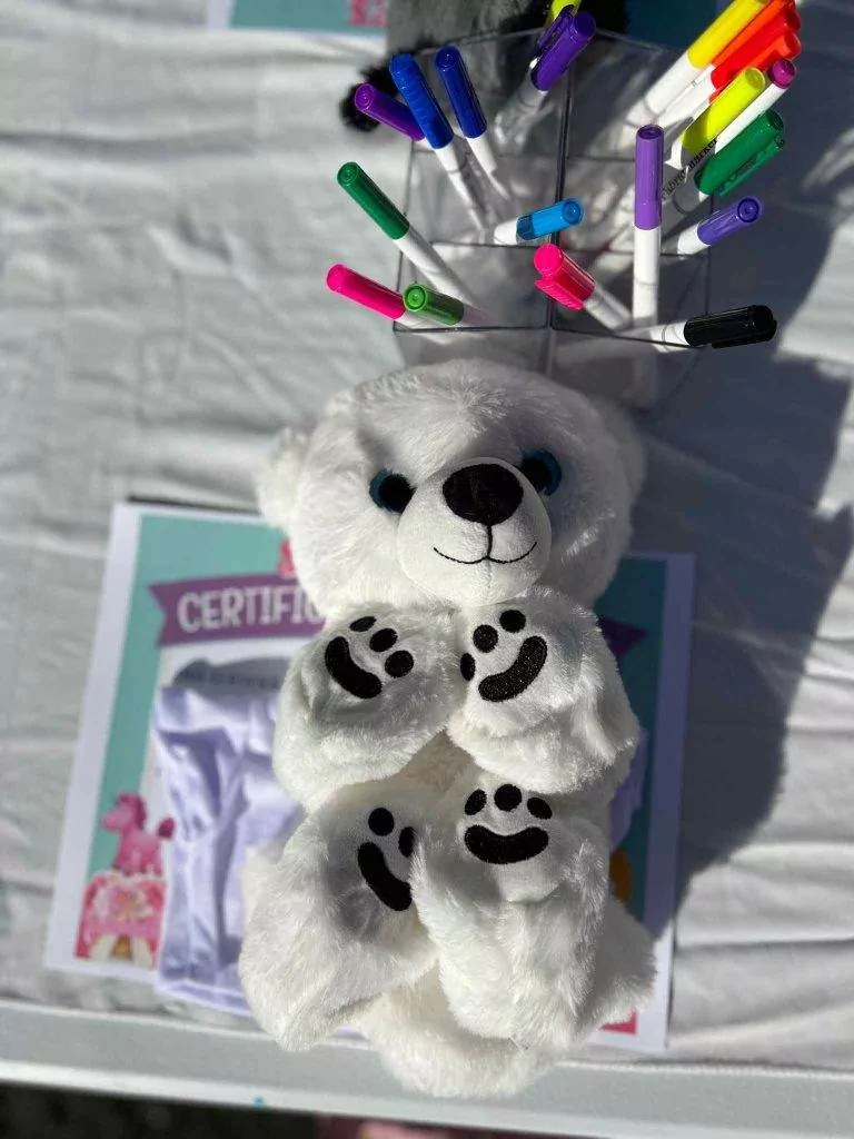 White teddy bear surrounded by colorful markers and a certificate on a table, set up for Build A Stuffed Animal Party activities, in bright outdoor lighting.