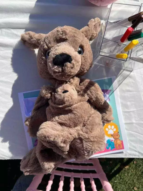 Two stuffed animal kangaroos, a mother and joey, sitting on a table with a children's book and pens in the background.