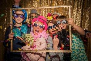 Five children at a slumber party wearing funny costumes and holding props, posing behind a decorative frame against a gold glitter backdrop.