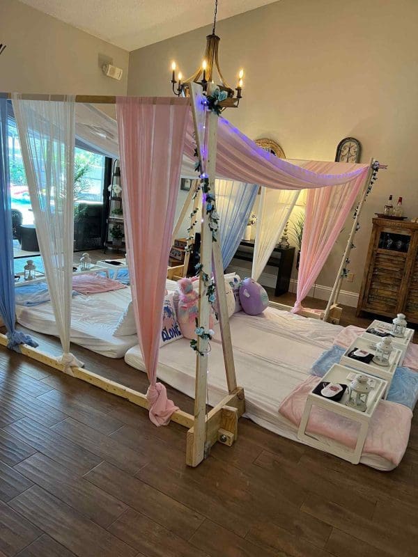 Indoor sleepover party setup with mattresses under a wooden frame draped with pink fabrics and fairy lights, surrounded by sheer curtains and cozy cushions.