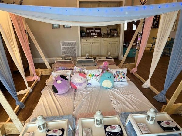 Indoor camping setup for kids, featuring canopy beds, plush toys, lanterns, and a decorative sign reading "Wavering Hole.