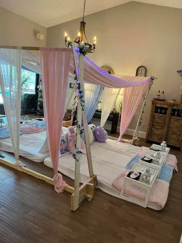 A cozy indoor bedroom setting with a canopied bed draped in pink curtains, adorned with fairy lights and ivy, next to a tray with spa essentials, perfect for a Canopy Slumber Party