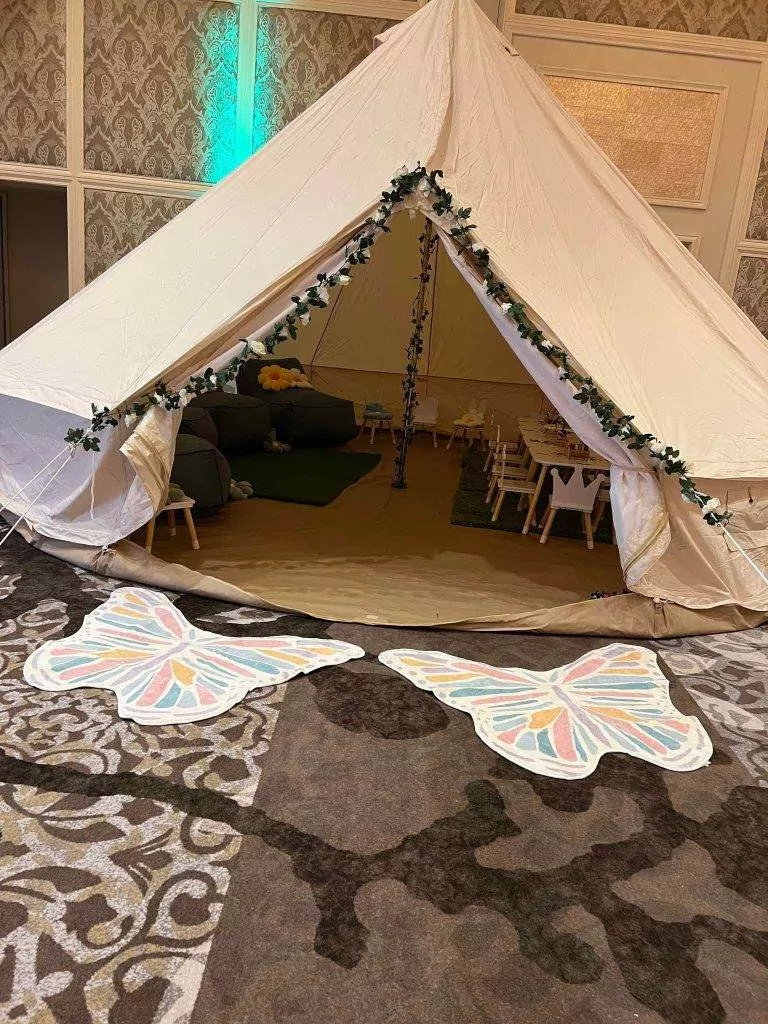 A large indoor tent decorated with leafy garlands at the entrance, flanked by two butterfly-shaped mats on a patterned carpet, perfect for various party themes.