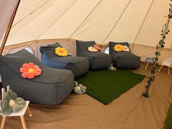 A cozy tent interior designed with party themes in mind, featuring gray bean bag chairs decorated with colorful flower cushions, set on a green rug and beige floor.