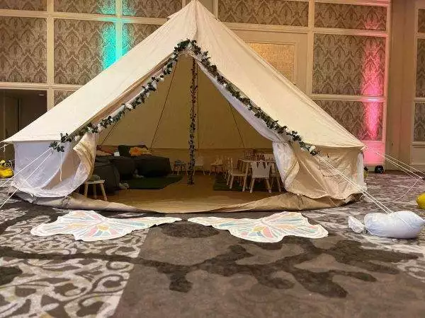 A large indoor tent themed for a party, decorated with fairy lights and foliage, furnished with a couch and small tables, set up on a patterned carpet.