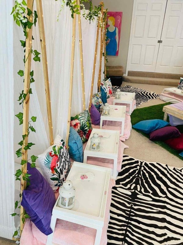 Indoor tropical birthday theme picnic setup with colorful cushions, small tables decorated with lanterns, and curtains draped over wooden frames.