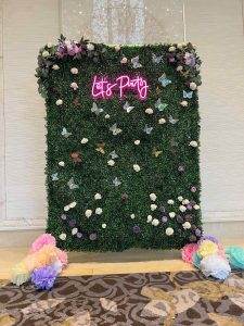 A decorative photo backdrop with a greenery wall adorned with colorful flowers and butterflies, featuring a neon sign that reads "Winter Haven FL Party.