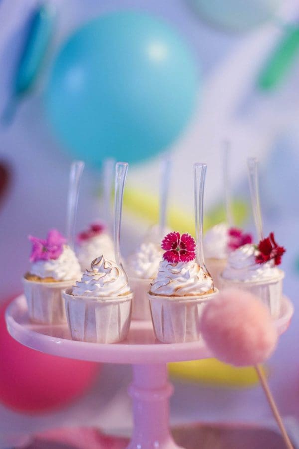 Mini cupcakes with whipped cream and pink flowers on top, displayed on a pink stand against a backdrop of colorful balloons for a tropical party theme.