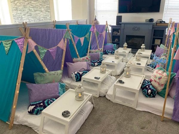 Indoor camping setup with magical teepees and lanterns placed on white tables in a living room, ready for a mermaid slumber party.