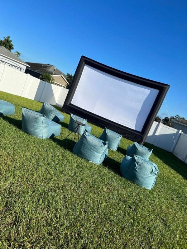 Outdoor movie setup with a large inflatable screen and several blue bean bag chairs on grass, ideal for a birthday party add-on, under a clear blue sky background.
