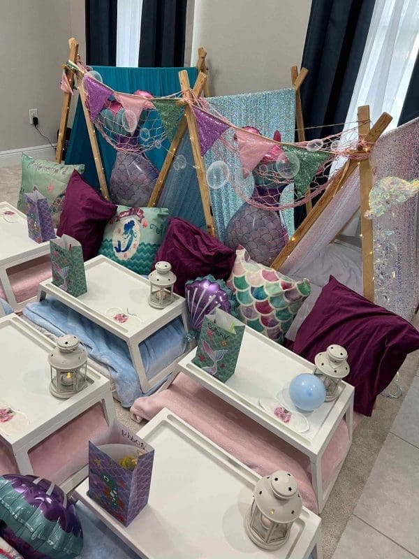 Indoor teepee fort decorated with mermaid-themed pillows, blankets, and lanterns, arranged around small white tables for a children's slumber party.