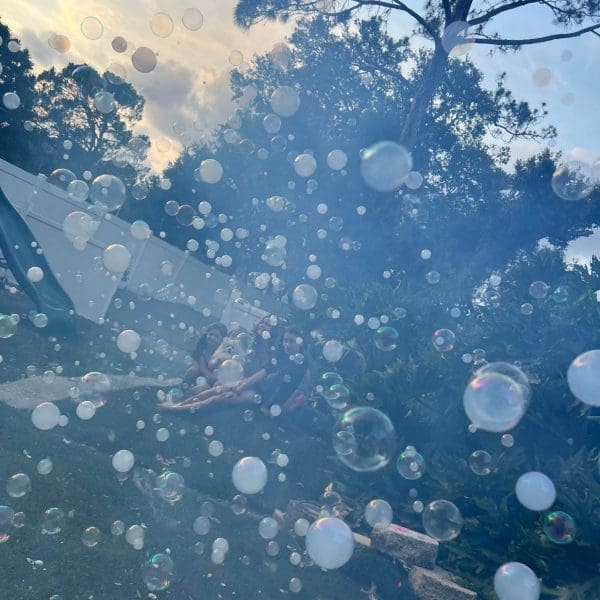 A hazy outdoor scene filled with numerous floating soap bubbles, serving as birthday party add ons, with blurred figures of people visible in the background under trees.