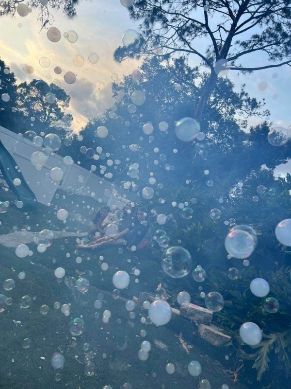 A hazy outdoor scene filled with numerous floating soap bubbles, serving as birthday party add ons, with blurred figures of people visible in the background under trees.