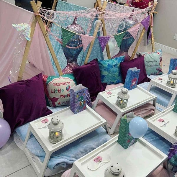 A mermaid slumber party setup with colorful teepees, pillows, and small tables decorated with lanterns and balloons.