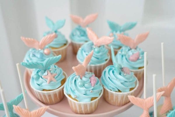 A tray of cupcakes decorated with blue frosting and topped with pink and blue fondant seashells and starfish, perfect for a Mermaid Slumber Party, on a pink background.