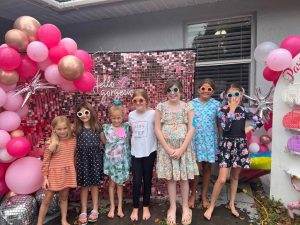 Group of seven children posing in front of a pink balloon-decorated backdrop with a sign saying "hello gorgeous" at a party themed event.