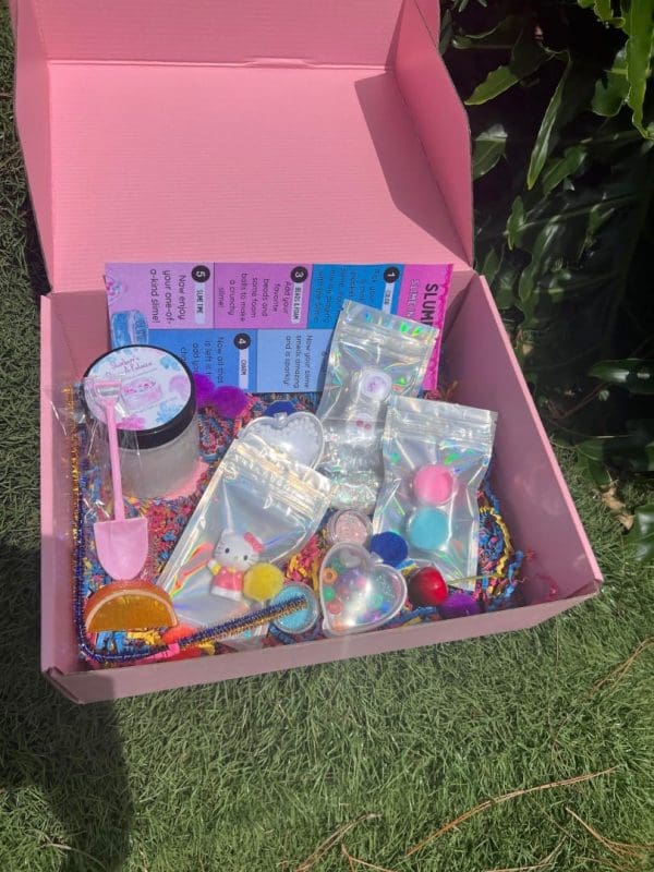 An open pink box containing various colorful children's toys and party favors, including bracelets, a shovel, and small containers, set on grass—delivered to your doorstep.