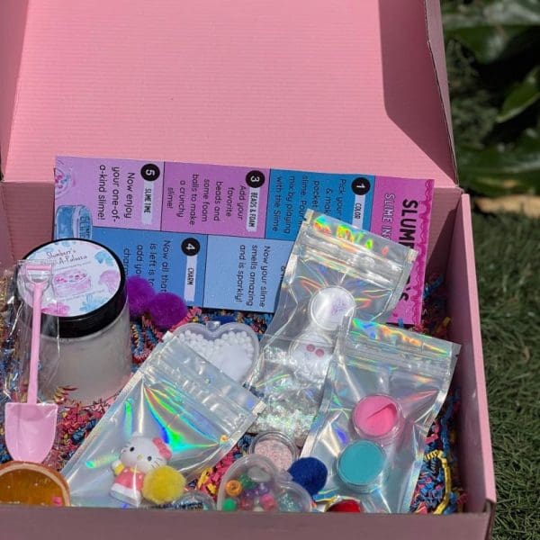 An open pink box filled with an assortment of crafting supplies, including beads, stickers, glitter, and small containers, placed on grass—fun delivered to your doorstep.