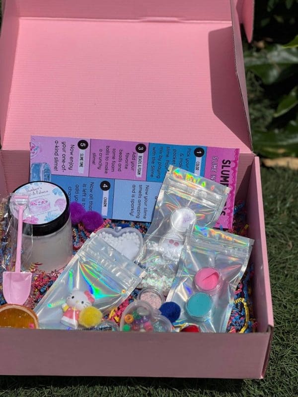 An open pink box filled with an assortment of crafting supplies, including beads, stickers, glitter, and small containers, placed on grass—fun delivered to your doorstep.