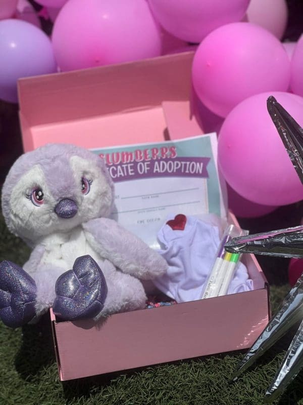 A plush toy koala in a pink Build A Stuffed Animal gift box with a certificate of adoption, surrounded by pink and purple balloons on grass.